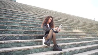 Kıvırcık saçlı şık bir kadın canlı bir şehirde açık havada merdivenlerde tabletini kullanıyor. Arkaplan, moda ve teknolojiyi harmanlayan rahat ve teknolojik yaşam tarzını ön plana çıkarıyor.