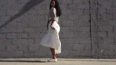 Kendine güvenen beyaz etekli ve desenli bluzlu bir kadın gri bir duvara karşı zarif bir duruş sergiliyor. Moda, zarafet ve sanatsal bir ifade sergiliyor. Güzelliği ve kişiliği yansıtıyor.