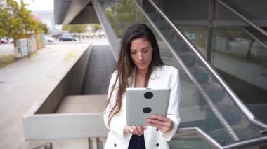 Modern bir binanın yakınında modern bir tablet kullanan şık ceketli profesyonel bir kadın, teknoloji ve üretkenlik karışımının başarılı kariyeri için çok önemli olduğunu gösteriyor.