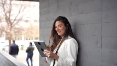 Modern gri bir duvarın önünde dijital tabletle uğraşan şık ve kendinden emin bir iş kadını, profesyonellik ve zarafetin mükemmel bir karışımını sergileyerek gülümsüyor.