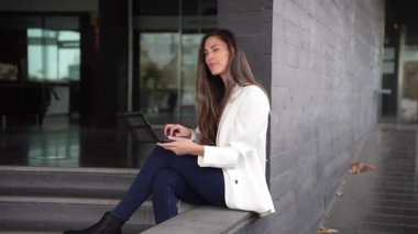 Uzun saçlı genç bir kadın, çalışanlar arasında yaratıcılığı, işbirliğini ve yenilikçi düşünceyi geliştirmek için tasarlanmış modern bir ofiste merdivenlerde otururken dizüstü bilgisayarına odaklanıyor.