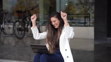 Neşeli genç bir kadın hoş bir havada dışarıda dizüstü bilgisayarını kullanarak neşe saçıyor. Bisikletler ve yeşilliklerin modern arkaplanı yaratıcılık ve işbirliği için üretken bir atmosferi teşvik etmektedir.