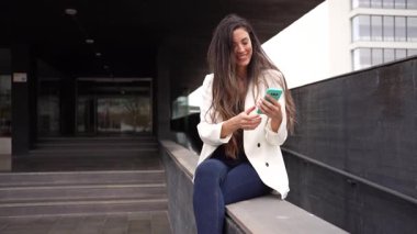 Neşeyle dolu genç bir kadın, şu anda kendisini çevreleyen canlı ve modern mimari manzaranın ortasında rahat bir şekilde otururken şık akıllı telefonuyla mesajlaşıyor.