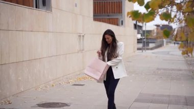 Güzel bir sonbahar gününde zarif, modern bir şehir parkında gezen şık bir genç kadın elinde renkli alışveriş torbalarıyla çarpıcı mevsimsel manzaranın tadını çıkarıyor.