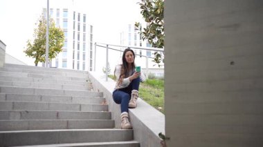 Genç bir kadın şık bir şehir binasının merdivenlerinde, modern mimari ve yemyeşil bir ortamda sabah kahvesini yudumlayarak huzurlu bir atmosfer yaratıyor.