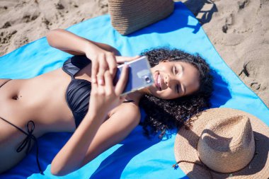Güneşli bir günde, genç bir kadın mavi plaj havlusuyla eğleniyor, neşeli bir selfie çekiyor. Hasır şapkası ve parıldayan kumu kaygısız plaj havasını arttırır. Unutulmaz anılar için mükemmeldir.