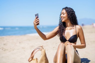 Şık bir siyah bikini giyen neşeli bir kadın, harika bir plaj gününün tadını çıkarırken aynı zamanda yaz atmosferinin ve onun hoş, kaygısız yaşam tarzının özünü mükemmel bir şekilde yansıtan neşeli bir selfie çekiyor.