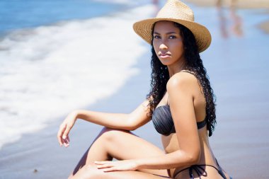 Bir manken şık bir bikini ve hasır şapkayla kumsalda oturur, güneşin tadını çıkarırken kendine güven yayar, mükemmel yaz atmosferini yakalar ve kaygısız sahil yaşam tarzını benimser.