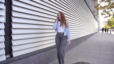 Dalgalı uzun saçlı genç bir kadın hareketli bir cadde boyunca güvenli bir şekilde geziniyor, telefonuyla canlı bir konuşma yapıyor, şık şehir tarzını parlak, canlı çizgili bir duvara karşı sergiliyor.