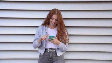 Uzun, dalgalı kızıl saçlı genç bir kadın, akıllı telefonuyla özümseyen ve günümüzün çağdaş şehir yaşam tarzını temsil eden canlı çizgili bir duvara yaslanıyor.