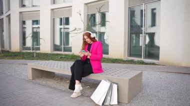 Kalabalık bir şehir ortamında, parlak pembe ceketli şık bir kadın bir bankta oturuyor, akıllı telefonuna odaklanmış, alışveriş torbalarıyla çevrili, dışarıda, canlı atmosferde günün tadını çıkarıyor.