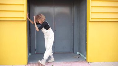 Kıvırcık saçlı enerjik genç bir kadın şık kıyafetini sergiliyor, bir şehir girişinde şakacı bir şekilde poz veriyor. Etrafındaki parlak renkler, sahnenin modern havasını ve sanatsal enerjisini arttırıyor.