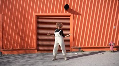 Şık ve şık bir dansçı, etkileyici ve büyüleyici hareketlerini ustalıkla sergiliyor. Arkasındaki çarpıcı turuncu kıvrımlı duvarı güzelce tamamlayan modaya uygun bir kıyafetle.