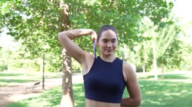 Güzel yeşil bir parkta, genç bir kadın direniş bandıyla çalışıyor, omuz germe hareketleri yapıyor. Açık hava ortamında aktif bir yaşam tarzını ve fitness terfisini temsil ediyor.