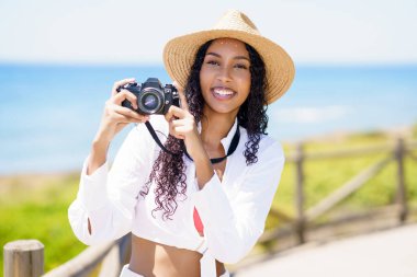 Şık bir şapkayla sahilde gülümseyen genç bir kadın, köpüklü suların ve yeşilliklerin ortasında, yaz eğlencesi ve maceranın somutlaştırıcısı, herkesi rahatlamaya ve neşeye davet ediyor.