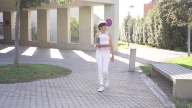 Kendinden emin genç bir insan, yemyeşil çimlerin modern mimariyle zemin üzerinde dans eden neşeli gölgelerle dolu güneşli bir günde, tezat oluşturduğu canlı bir şehir parkında gururla duruyor.