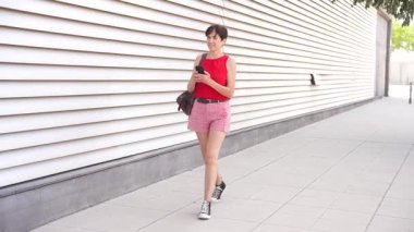 Parlak kırmızı bir bluz giyen sıradan bir genç kadın şık bir ekose şortla modern bir şehir sokağında yürürken akıllı telefonuna odaklanıyor ve ekranında ne varsa onunla ilgileniyor.