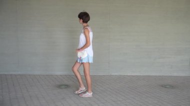 Genç bir kadın şık bir yaz kıyafetiyle, basit, sade bir arka planla tezat kurarak, özgüven ve gayret sarf ederek, hareketli bir sokakta yürüyor.