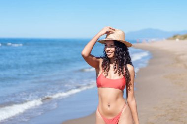 Güzel bir kadın güneşli bir sahilde neşe saçar, şık bir bikini ve zarif bir güneş şapkası giyer. Bu sahne eğlence ve rahatlama ruhunu yakalar, yaz temalı projeler için mükemmeldir.