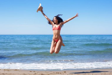 Parlak pembe bir bikini giyen neşeli bir kadın kumlu sahilde neşeyle sıçrayıp yaz eğlencesinin, heyecanın ve güzel okyanusun yanında özgürlüğün gerçek özünü yakalıyor.