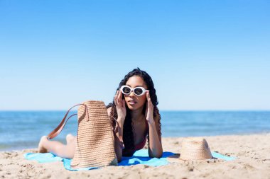 Şık genç bir kadın plajda dinleniyor, şık güneş gözlükleri ve renkli aksesuarlarıyla şık görünüyor, kaygısız doğasıyla yaz titreşimlerini somutlaştırıyor, güneşin altında eğleniyor.