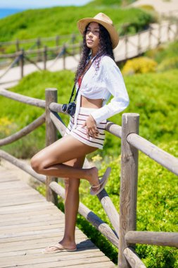 Genç bir kadın, şık yaz kıyafetlerini ve modaya uygun hasır şapkasını sergileyerek, güzel tasarlanmış ahşap bir yolda güvenli bir şekilde poz veriyor, canlı yeşil manzaranın ortasında hem keyif hem de zarafeti somutlaştırıyor.