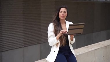 Şık beyaz ceketli şık bir kadın, modern yaşam tarzını ve çalışma ahlakını ön plana çıkaran çağdaş şehir zeminine karşı tabletine odaklanmış modaya uygun bir yerde oturuyor.