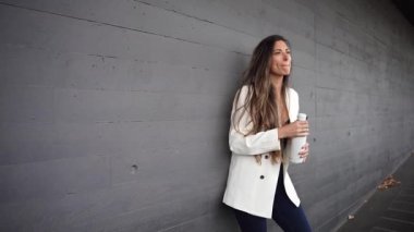 Uzun, dalgalı saçları olan şık genç bir kadın güzel dokulu bir duvara yaslanır, sıcak kahvesini yudumlar ve huzur dolu bir anın tadını çıkarır.