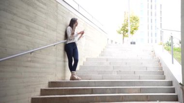 Genç bir kadın, modern bir kentsel ortamda, kendine güvenen bir set beton merdivende dikilirken, çevresindeki sesleri ve manzaraları seyrediyor.