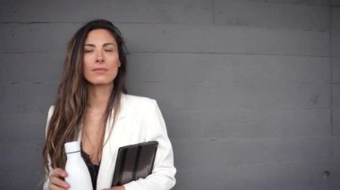 Uzun ve dalgalı saçları olan bir kadın modern çağdaş bir ortamda derin bir düşünceli bir hava yayarken elinde telefonla şık ve şık bir gri duvara yaslanıyor.