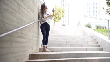 Genç bir kadın, dijital dünyasına dalmış, canlı ve modern bir şehir ortamının merdivenlerinde otururken akıllı telefonunu dikkatle kullanarak beton bir duvara yaslanıyor.