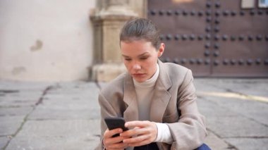 Kendini işine adamış ve odaklanmış profesyonel bir kadın, akıllı telefonuyla açık hava ortamında rahat bir şekilde yerde oturuyor ve modern dijital iletişimin özünü vurguluyor.