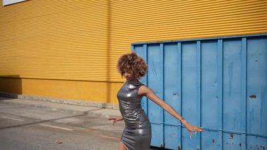 Şık bir kadın canlı mavi bir duvarın yanında, canlı sarı kıvrımların göz alıcı zeminine yaslanmış, dinamik kıyafetiyle çağdaş şehir modasını sergiliyor.