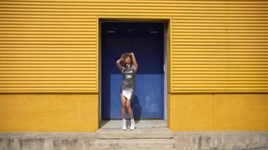 Şık, genç bir kadın, neşeli, sarı bir duvarın önünde canlı enerjisini ve neşeli kişiliğini yansıtan modaya uygun kıyafetlerini sergileyerek rahatça poz veriyor.