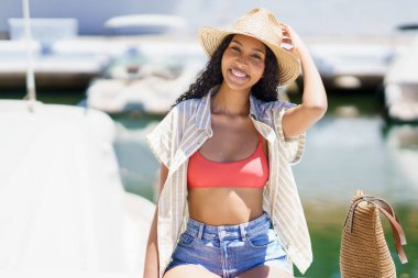 Neşeli genç bir kadın zarif bir şekilde güzel bir marinanın yanında poz veriyor, şık kıyafetinin içinde canlı yaz titreşimleri yayıyor. Bu sahne plaj kıyafetleri kampanyaları ya da sezonluk tanıtım etkinlikleri için mükemmel.