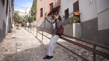 Tarz sahibi genç bir kadın tesadüfen bir korkuluğun tepesinde oturur, büyüleyici bir kaldırım taşı caddesinde neşeli bir selfie çeker. Etrafı büyüleyici kırsal binalarla çevrilidir, mükemmel bir şekilde biçimlendirilmiş moda ve eğlenceyle doludur.