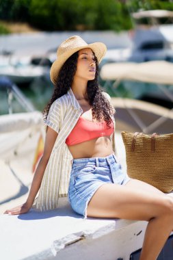 Şık ve modaya uygun genç bir kadın, şık bir mayo ve şık bir hasır şapka giyerek, kendine güveni, mutluluğu ve rahatlama duygusuyla marinada güzel bir yazın tadını çıkarıyor.