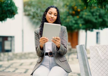 Moda sahibi bir kadın dışarıda oturuyor, tabletine dalmış, etrafını alışveriş torbaları sarmış, modern şehir yaşam tarzını ve teknisyenliğini mükemmel bir şekilde somutlaştırıyor.