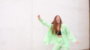 Uzun kızıl saçlı neşeli bir kadın yeşil bir kıyafetle beyaz bir duvara karşı enerjik bir şekilde dans ediyor. Mutluluk ve canlılık saçıyor. Dinamik enerjiyle dolu kaygısız bir titreşim yayıyor.