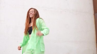Neşeli, şık, kızıl saçlı bir kadın, enerjik bir şekilde dışarıda dans ediyor, canlı ve tasasız bir enerji saçıyor, modaya uygun yeşil bir elbise içinde, modern, dinamik ve anlamlı hareketler sergiliyor.