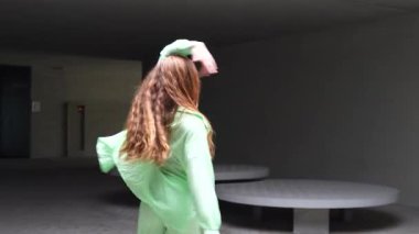 Neon yeşil elbiseli genç bir kadın modern bir kapalı alanda neşeyle dans ediyor. Dinamik hareketleri yaratıcılığı ve canlılığı gösteriyor, özgür bireyselliği ve canlı bir ruhu ifade ediyor.