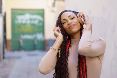 Saçları güzel örülmüş genç bir kadın neşeli bir şekilde en sevdiği müziği dinlerken açık havanın tadını çıkarıyor. Etrafında renkli arka plana yeni bir renk katan hareketli bir şehir manzarası var.