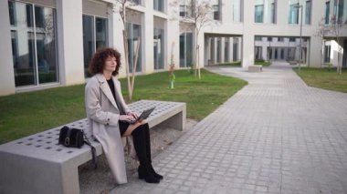 Profesyonel bir kadın açık havada şık bir bankta oturur, laptopuna odaklanır, etkileyici modern mimarinin ortasında iş ve doğa arasındaki uyumu canlı ortamında gösterir.