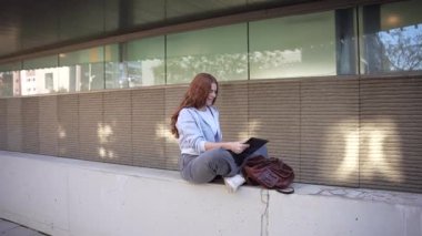 Genç bir kadın, büyüleyici bir kitaba gömülmüş modern bir duvarda dinleniyor. Onun rahat duruşu, çağdaş bir şehirde şık ve neşeli bir yaşam tarzını yansıtan canlı şehir zeminiyle harmanlanır.