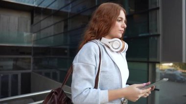 Tabletine kendini kaptırmış trend bir genç kadın, modern çağın ve profesyonellerin modern yaşam tarzını temsil eden, bağlantı ve yenilikçiliği somutlaştıran parlak bir cam zemin karşısında duruyor.