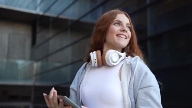 Güzel kıvırcık saçlı genç bir kadın, tablet bir cihaz kullanırken, şık kulaklıklar takarken, parlak bir şekilde gülümsüyor ve çağdaş ve canlı bir ortamda dışarıda keyifli bir anın tadını çıkarıyor.