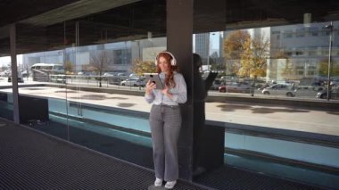 Gündelik ama şık bir şekilde giyinmiş genç bir kadın, kendisini çevreleyen hareketli ve dinamik modern şehir manzarasına tamamen dalmış, akıllı telefonuna tamamen dalmış, bir sütunun karşısında rahat duruyor.