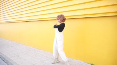 Modaya uygun genç bir kadın şık tulumuyla canlı sarı bir duvara karşı neşeyle dans ediyor. Sokak tarzının özünü her hareketinde ve kıyafetinin ifadesinde yakalıyor.