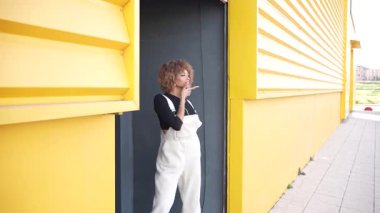 Güzel, kıvırcık saçlı şık bir genç kadın şık beyaz tulumlar içinde canlı ve canlı sarı bir arkaplan karşısında zarifçe poz verir. Kendine güvenen bir hava yayar ve yaratıcılığını sergiler.