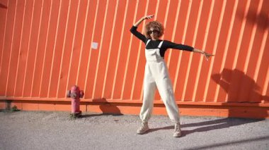 Modayı takip eden genç bir kadın, şık bir tulum ve parlak siyah bir bluz giyiyor, canlı turuncu bir duvara karşı kendine güvenen bir poz veriyor, kentsel moda ve yaratıcılığın özünü vurguluyor.